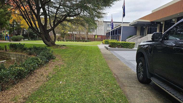 Freshly mown lawn and clipped garden beds beside the paved entrance path of a club building with flagpoles