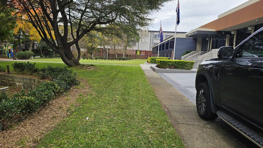 Freshly mown lawn and clipped garden beds beside the paved entrance path of a club building with flagpoles
